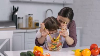 Sevgi dolu bir anne ve gülümseyen küçük kızı mutfakta sebze salatası karıştırma eşleşen önlük giyen özel ihtiyaçları olan. Down sendromlu anneler yardım ile yemek öğrenme neşeli kız.