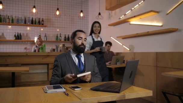 可爱的混血女服务员在舒适的咖啡店里为认真的胡须商人端上一杯热咖啡 成功的男性企业家在咖啡馆加班 喝热饮 — 图库视频影像