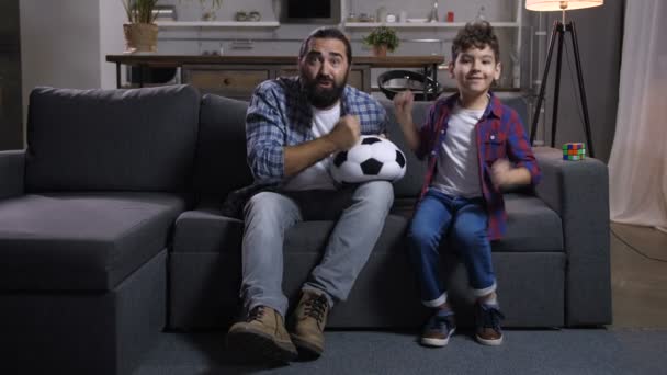 兴奋的父亲和混血的儿子在电视上看体育比赛 欢呼足球队 失望的爸爸与孩子表达负面情绪后 错过了一个进球 在足球游戏广播在电视上 — 图库视频影像