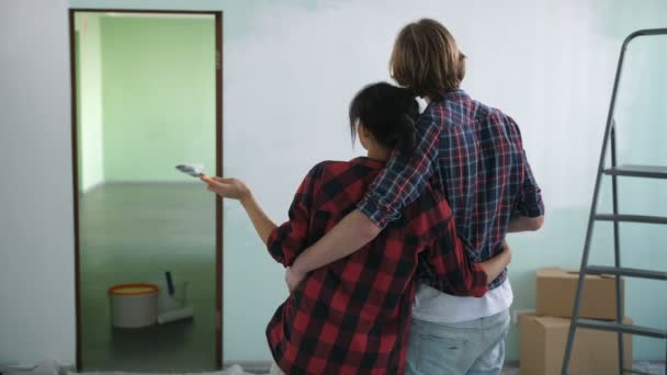 Rear view of renovation diy couple with paintbrush and paint roller embracing and looking at half painted wall in new house. Creative couple discussing idea of new interior design during home makeover — Stock Footage Rear View Renovation Diy Couple Paintbrush Paint Roller Embracing Looking Royalty Free Stock Video