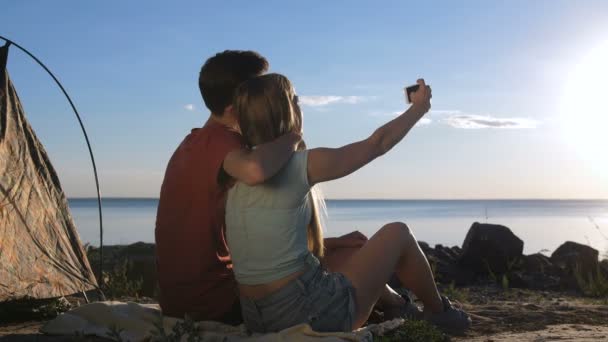 キャンプテントの近くの海岸に座って 夏のキャンプ旅行中に美しい海の景色を楽しみながら スマートフォンでセルフポートレート写真を作る幸せな若いカップル — ストック動画