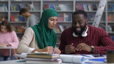 Arka planda öğrenme bulanık beyaz üniversite öğrencileri ile Kitaplığı'nda eğitim sırasında pozitif Afro-Amerikan erkek öğrenciye yeni taslak proje açıklayan hijab güzel Müslüman kadın.