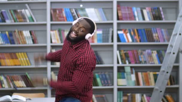 Homem negro alegre em fones de ouvido dançando na biblioteca — Vídeo de Stock