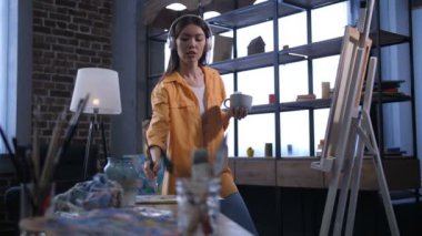 Onu eve art Studio tuval üzerine resim oluşturulurken kulaklık müzik dinleme kahve fincan ile neşeli profesyonel kadın sanatçı. İlham kadın ressam çizim resim atölyesi.