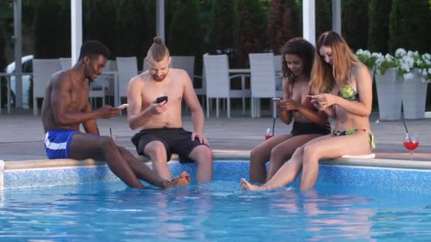 Blijde vrienden met behulp van telefoons zittend op zwembadzijde — Stockvideo