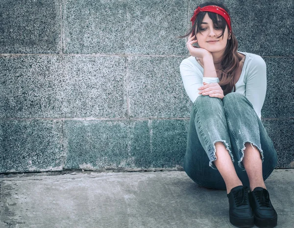 Beautiful girl in jeans and a white T-shirt with a red armband on the head, against the wall background, the concept of urban clothing and youth style