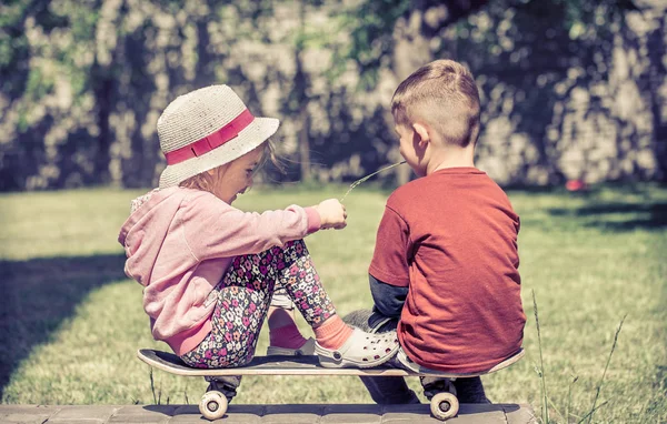 Menina e menino brincando no skate, contra o pano de fundo de um jardim verde, conceito de amizade infantil — Fotografia de Stock