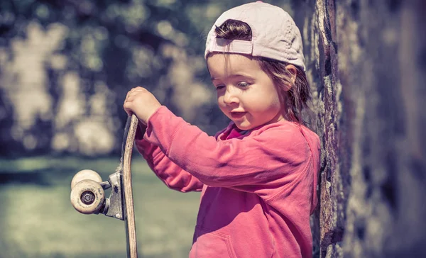 Ein modisches kleines Mädchen hält ein Skateboard in der Hand und spielt draußen, die schönen Gefühle eines Kindes. — Stockfoto