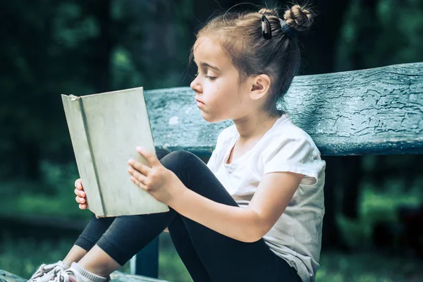 Маленькая Милая Девочка Сидит Скамейке Читает Книгу Парке Фоне Зеленой — стоковое фото