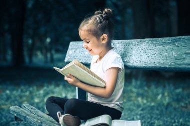 küçük sevimli kız bir bankta oturan ve parkta yeşil doğanın bir arka plan üzerine bir kitap okuma