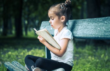 küçük sevimli kız bir bankta oturan ve parkta yeşil doğanın bir arka plan üzerine bir kitap okuma