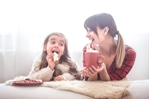 Mom and her cute daughter are eating Christmas sweets in the living room on a bright background, the concept of family values and festive atmosphere