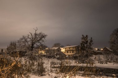 Kış gecesi kırsal üzerinde güzel kar. Ağaçlar, evler, yollar ve ışıklar ile kar sokak görünümü.