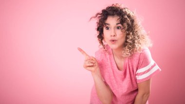 Gevşek pembe tişört kıvırcık saçlı Genç sürpriz kız portresi parmak pembe arka plan, metin, duygu kavramı için yer üzerinde izole tarafa gösterir
