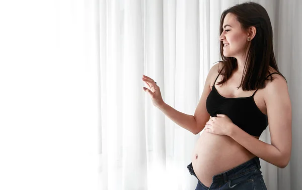 年轻的孕妇穿着短上衣 肚子和牛仔裤都很开放 站在家里的窗户附近 有做母亲的概念 也在期待孩子的出生 — 图库照片