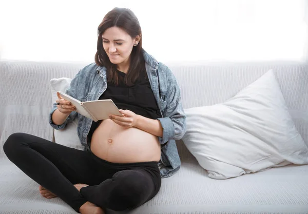 年轻美丽的孕妇坐在沙发上 在家里的窗边看书 怀孕的概念 期待着孩子的到来 — 图库照片