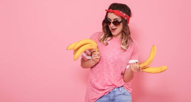 Güneş gözlüklü, şık giysiler içinde hipster kızla muz ellerinde, pembe bir arka plan, yaşam kavramı üzerinde yatay portresi