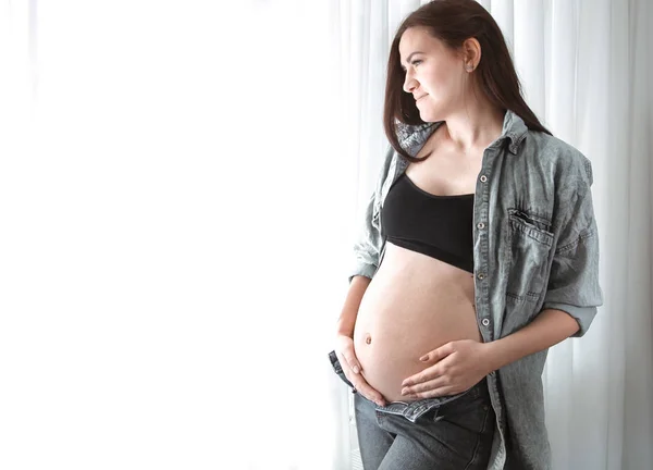 年轻的孕妇穿着短上衣 肚子和牛仔裤都很开放 站在家里的窗户附近 有做母亲的概念 也在期待孩子的出生 — 图库照片