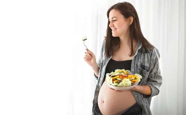 年轻孕妇在家里吃蔬菜沙拉附近的窗口 怀孕和健康饮食的概念 — 图库照片