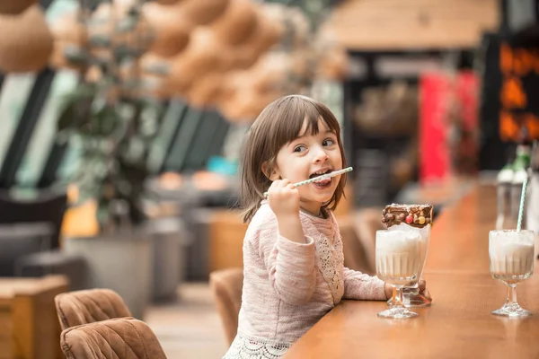 Charming Funny little girl drinks a milkshake in a restaurant, licking a tube. Baby food fast food . Leisure in the cafe.