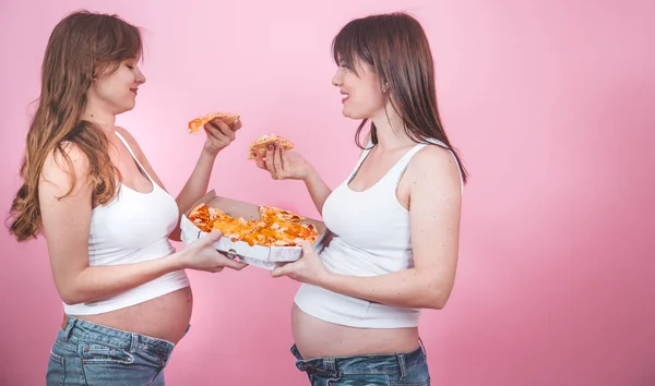 食物概念 两个美丽的孕妇吃比萨饼粉红色的背景 在牛仔裤和白色 女性友谊 — 图库照片