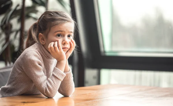 迷人的小女孩坐在窗边的一张木桌旁 心烦意乱的孩子 — 图库照片
