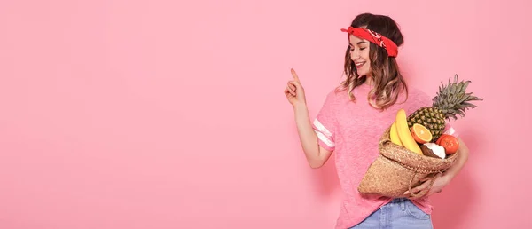 Красивая молодая девушка в розовой футболке, держит полный соломенный мешок фруктов на розовом фоне — стоковое фото