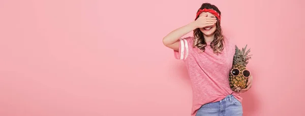 Красивая молодая девушка в розовой футболке, прикрывает лицо, держит ананас — стоковое фото