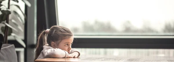 Γοητευτικό μικρό κορίτσι που κάθεται σε ένα ξύλινο τραπέζι από το παράθυρο. — Φωτογραφία Αρχείου