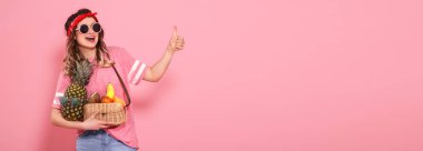 Pembe t-shirt ve gözlük güzel genç kız, pembe arka planda meyve tam bir saman çantası tutar. Finger sınıfını gösterir