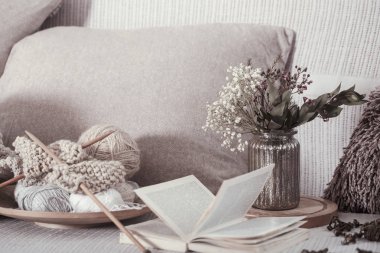 Vintage wooden knitting needles and threads on a cozy sofa with pillows and a vase of flowers. Open book for reading clipart