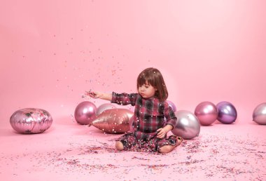 Pembe bir arka plan üzerinde oturan komik çocuk. Küçük kız balonlar ve konfeti ile eğleniyor