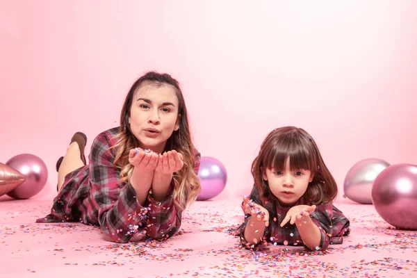 Funny mom and child lie on a pink background. Little girl and mother having fun with confetti