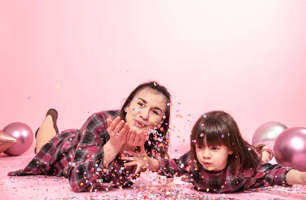 Funny mom and child lie on a pink background. Little girl and mother having fun with confetti