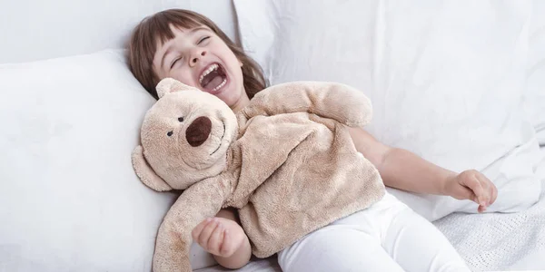 Süßes kleines Mädchen lächelt, während es in einem gemütlichen weißen Bett liegt — Stockfoto