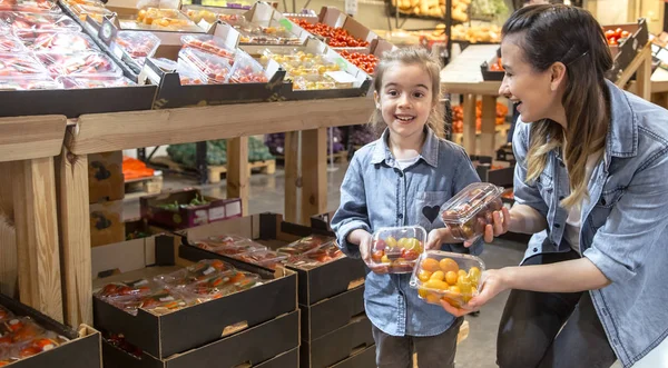 Cheerful smiling young woman with little daughter buying globe tomatoes at the market — Stock Photo, Image