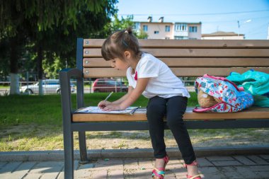 Sırt çantası olan bir ilkokul öğrencisi bir bankta oturuyor ve bir şeyler yazıyor..