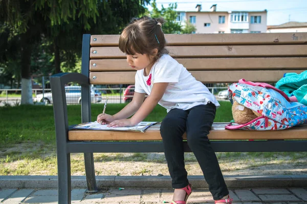 Sırt çantası olan bir ilkokul öğrencisi bir bankta oturuyor ve bir şeyler yazıyor.. — Stok fotoğraf