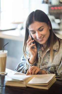 Bir kafede oturup kitap okuyan ve telefonda konuşan genç kız