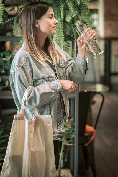 Niña bebiendo agua de una botella de vidrio con una bolsa ecológica en el fondo de macetas — Foto de Stock