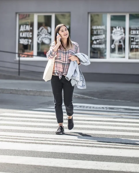 Chica joven cruzando la calle en un cruce peatonal con eco bolsa y teléfono — Foto de Stock