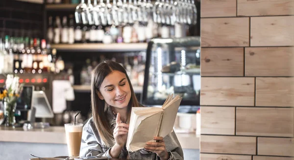 Niña sentada en un café leyendo un libro y bebiendo café con leche — Foto de Stock