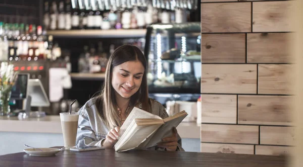 Niña sentada en un café leyendo un libro y bebiendo café con leche — Foto de Stock