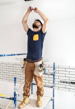 Elektrik İmalathanesi, yükseklerde lamba montajı. Profesyonel, tulumlu ve elektrikli aletli. Tamir alanının arka planında. Profesyonel olarak çalışma kavramı..