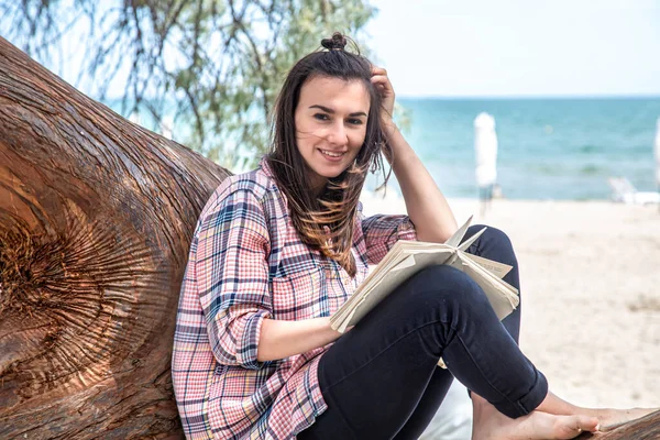 A happy girl reads a book on a tree, against the background of a sea beach. The person abstracted from everything. The concept of relaxation and tranquility.