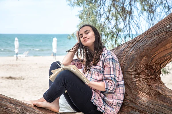 A happy girl reads a book on a tree, against the background of a sea beach. The person abstracted from everything. The concept of relaxation and tranquility.