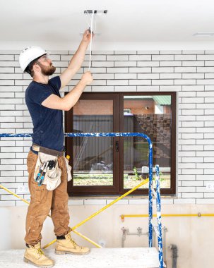 Elektrik İmalathanesi, yükseklerde lamba montajı. Profesyonel, tulumlu ve elektrikli aletli. Tamir alanının arka planında. Profesyonel olarak çalışma kavramı..