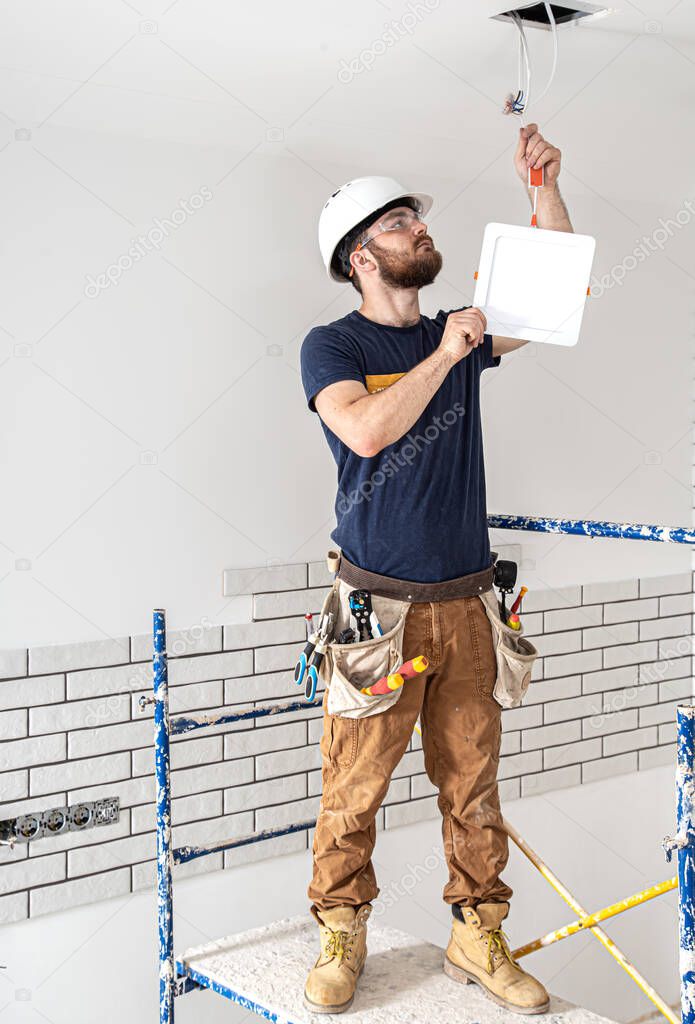Electrician Builder at work, installation of lamps at height. Professional in overalls with an electrical tool. On the background of the repair site. The concept of working as a professional.
