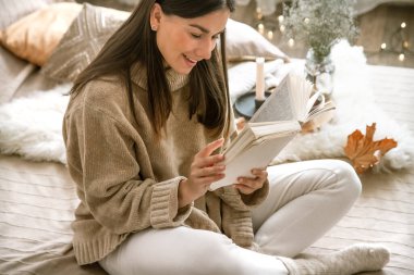 Süveter giyen genç bir kadın rahat bir evde kitap okuyor. Konfor ve hobi kavramı.