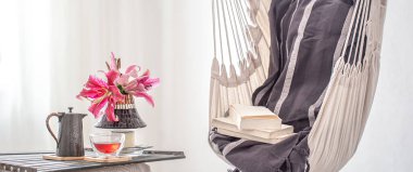 A boho-style hammock chair with a stack of books. Wooden tray with teapot and Cup of tea and beautiful Lily flowers. Cozy interior. The concept of rest and home comfort. Space for text. clipart